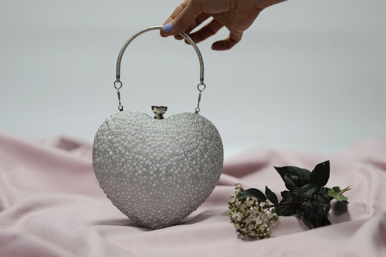 Stylish heart-shaped handbag held by a woman's hand with floral decor on a soft pink fabric background.